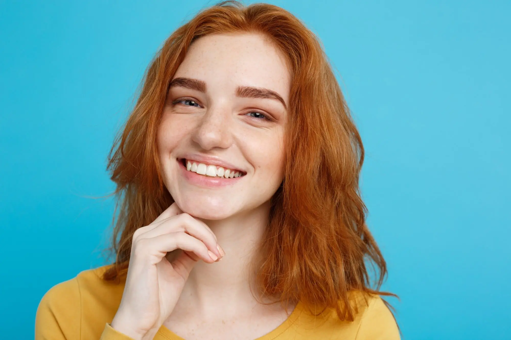 Woman with auburn hair smiles, resting a hand under her chin against a blue background in Sammamish, WA.