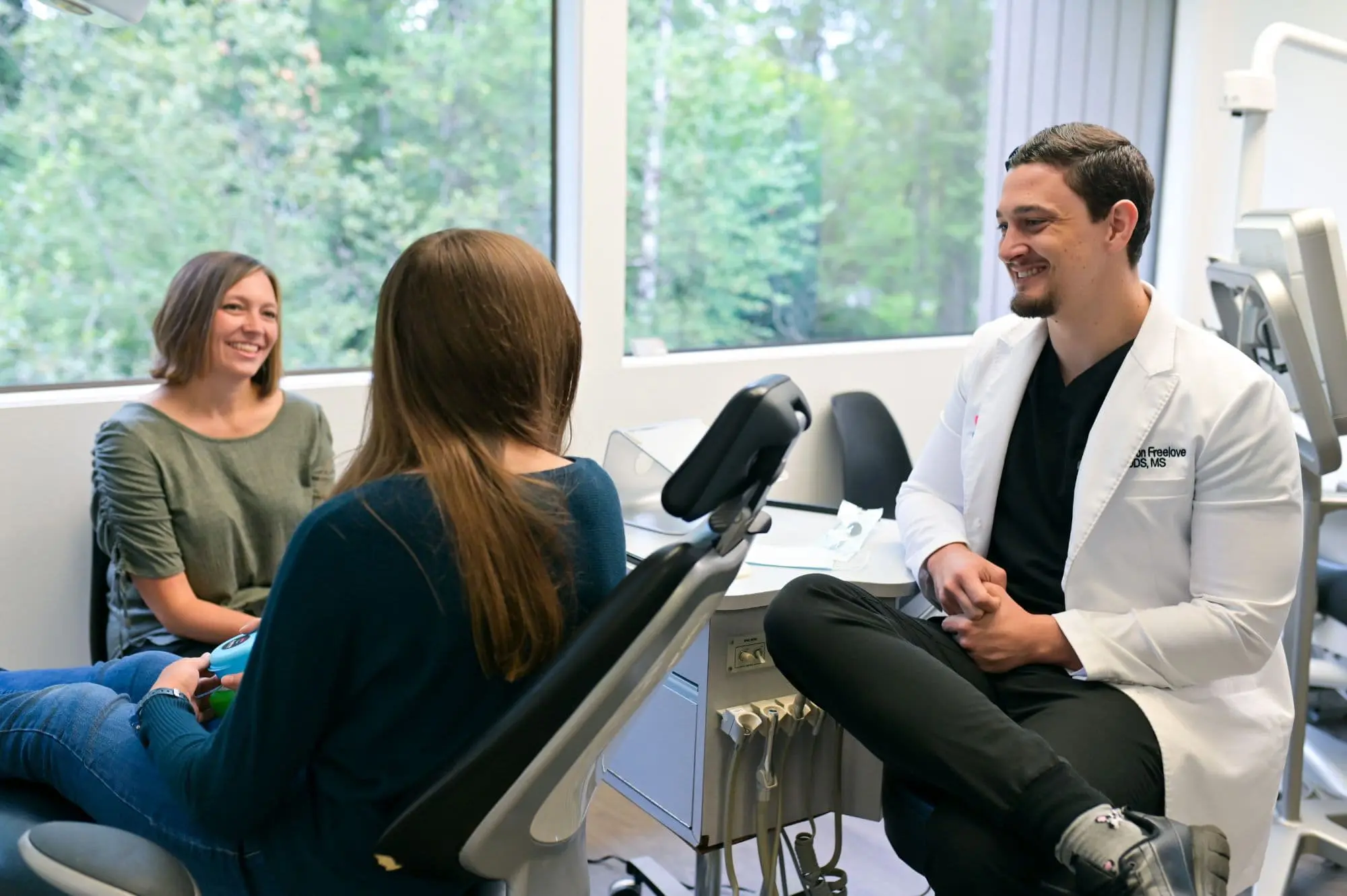 A dentist in a white coat discusses orthodontic insurance plans with two seated women in a dental office, where large windows reveal the lush greenery outside in Sammamish or Kent, WA