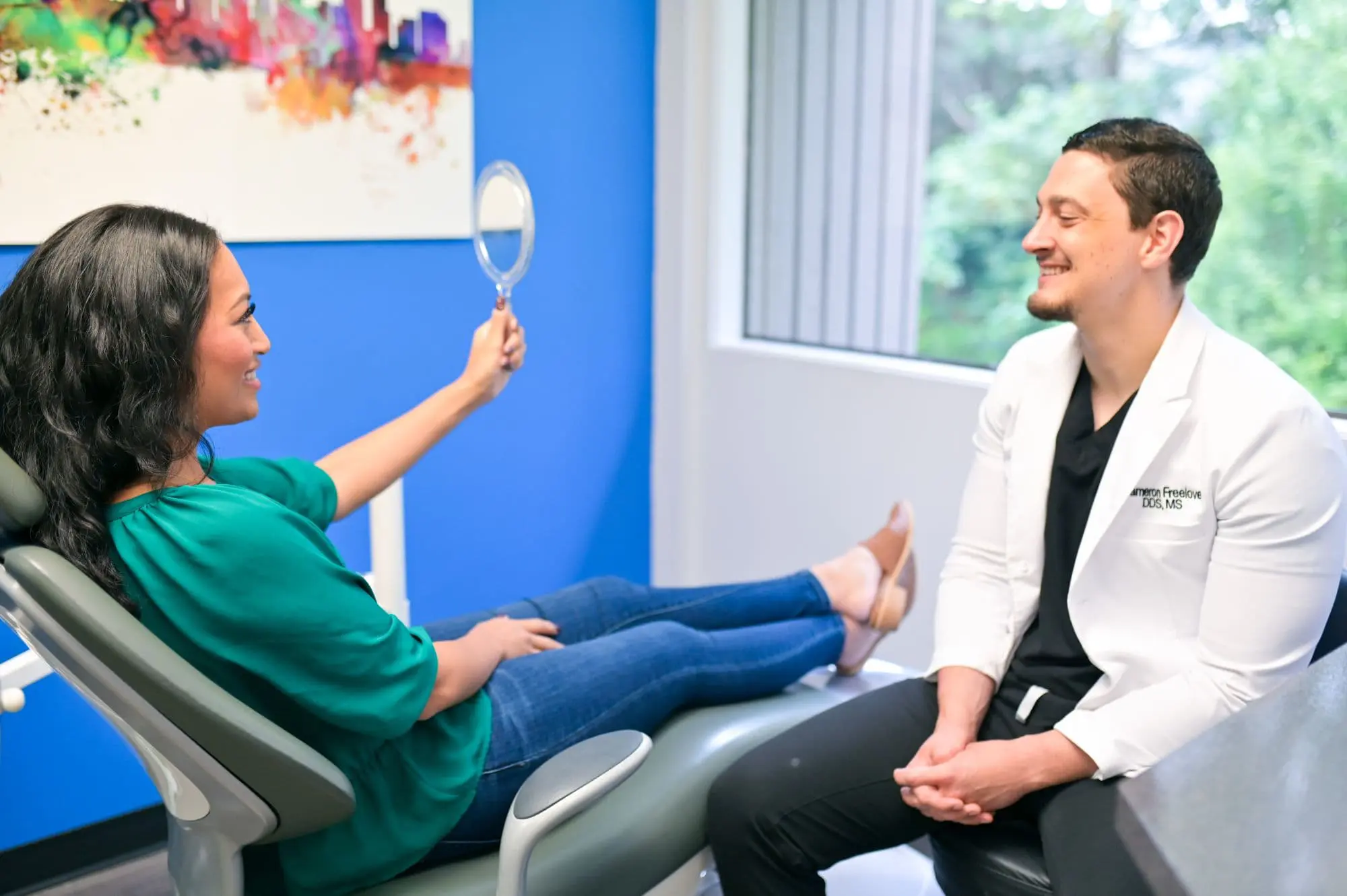 A woman with an underbite sits in a dental chair, holding a mirror and smiling at the dentist in a white coat beside her in the brightly lit room in Kent or Sammamish, WA