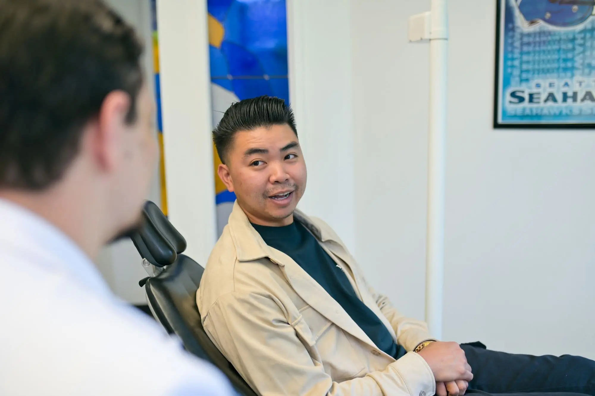 A person sits in a chair, focused on another who is slightly blurred, perhaps discussing orthodontic appliances. In the background, a Seahawks poster adds a pop of color to the scene in Kent, WA