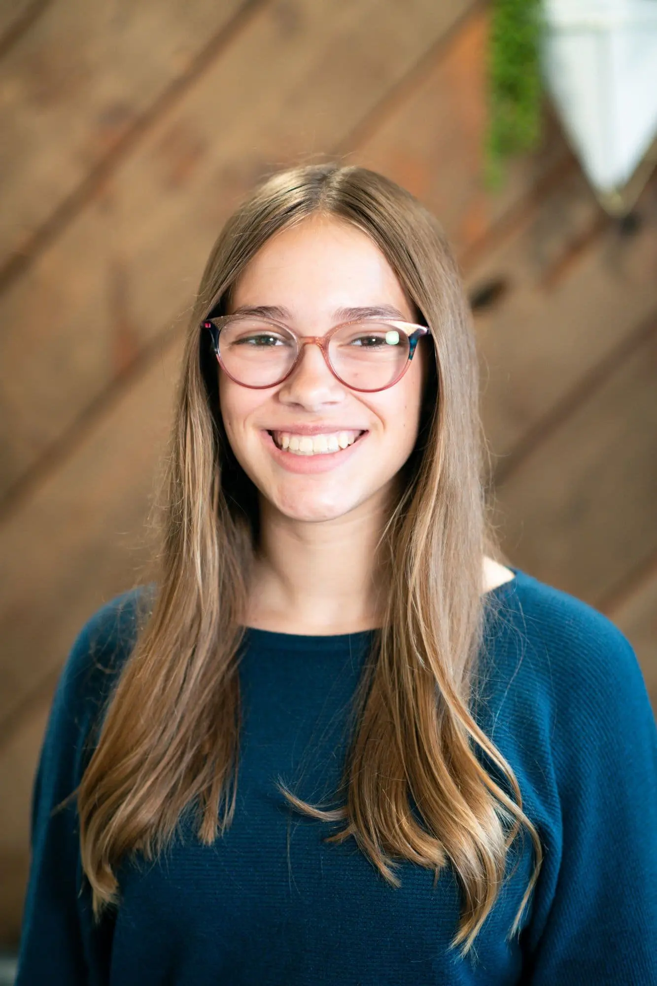 A person with long hair, glasses, and a charming overbite smiles warmly in a teal sweater. The scene is set against a wooden wall accented by lush greenery in Sammamish or Kent, WA