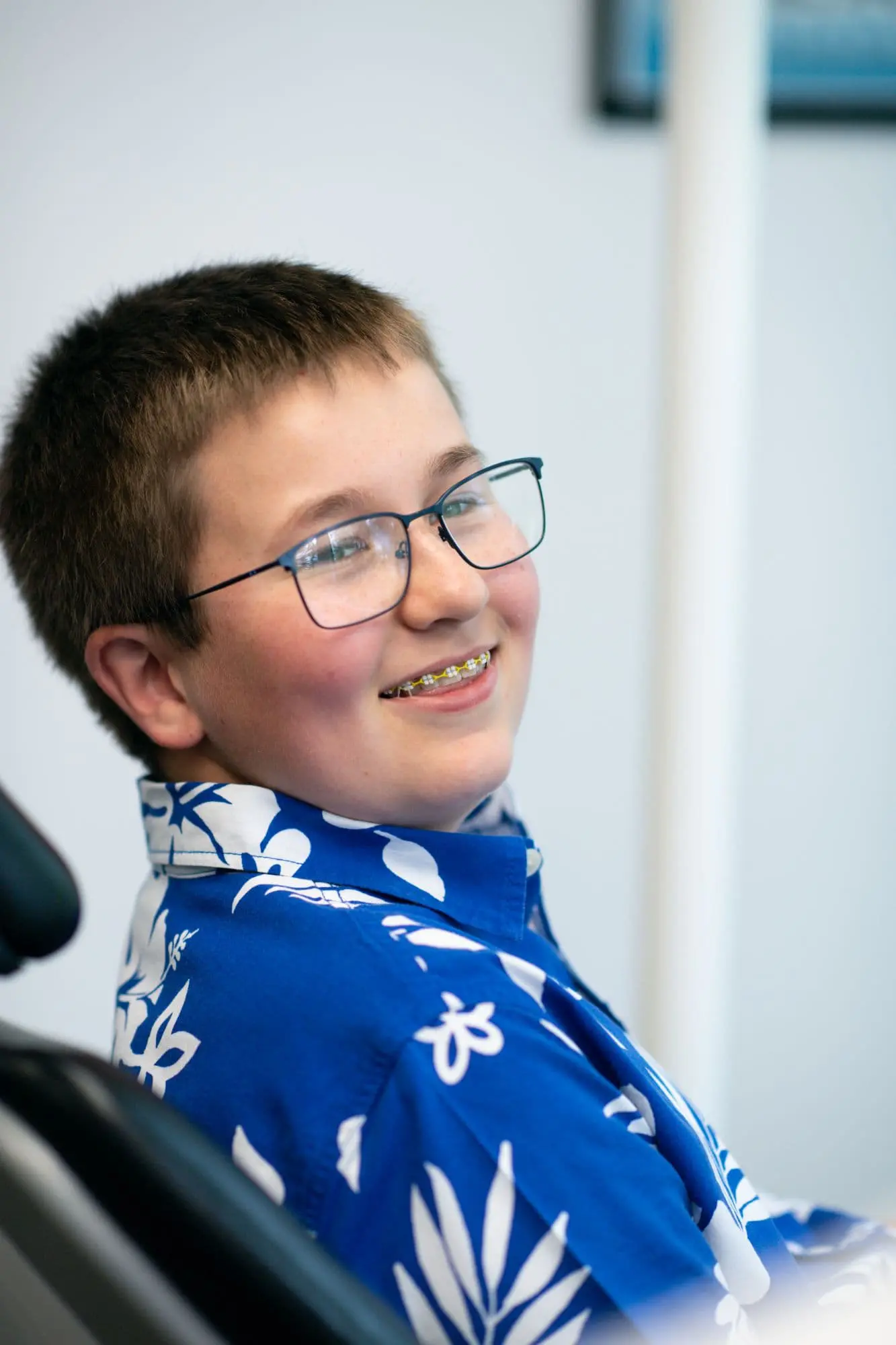 A smiling boy with braces and glasses dons a blue floral shirt, sitting comfortably as he looks towards the camera in Sammamish or Kent, WA