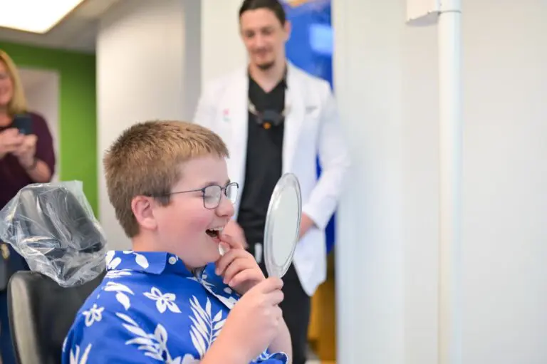 A boy in a floral shirt sits in a chair, smiling and inspecting his Invisalign in a hand mirror. A person in a white coat stands in the background, perhaps ready to discuss how treatment like this can be covered by insurance in Sammamish or Kent, WA