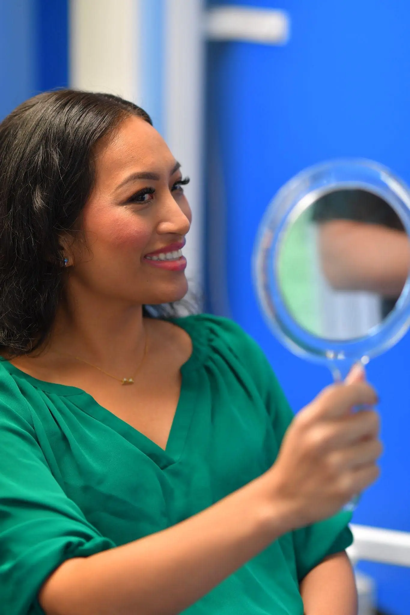 A woman in a green top smiles after dental braces treatment at her reflection in a handheld mirror in Sammamish, WA.