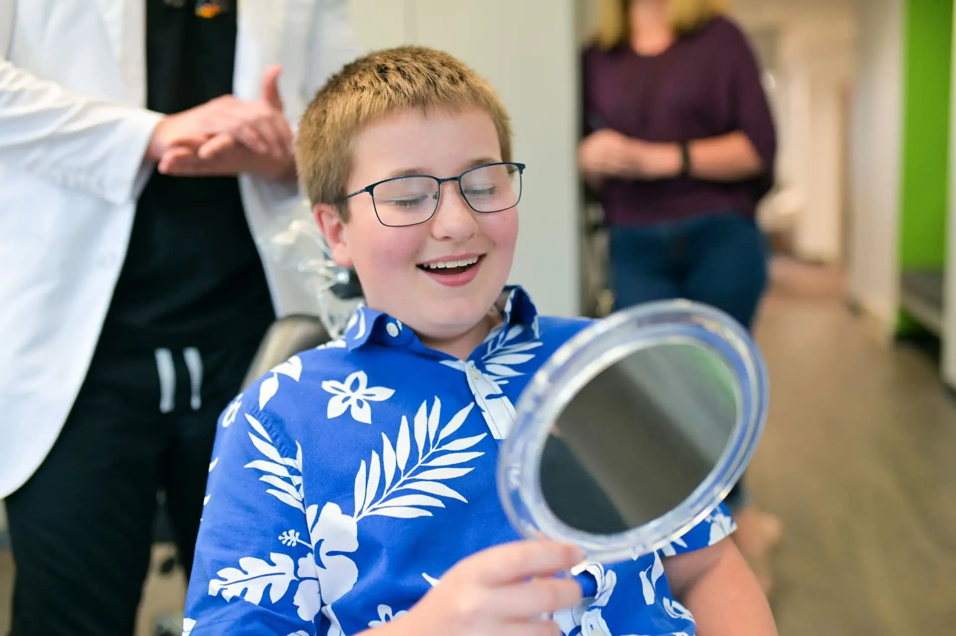 A boy wearing glasses and a blue floral shirt smiles while looking at a round mirror after metal braces treatment at Freelove Orthodontics in Kent, WA