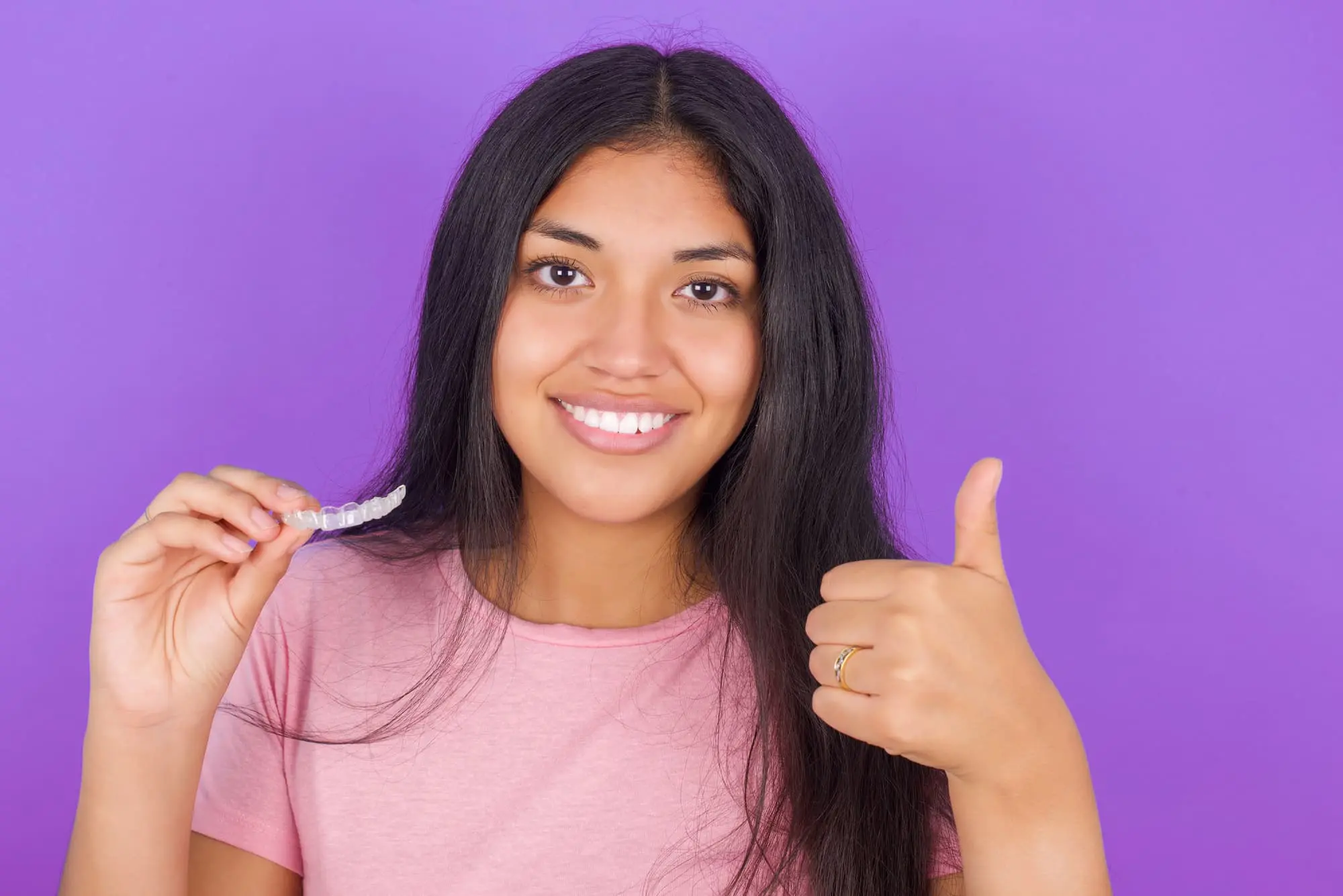 A woman with long dark hair holds affordable cheap Invisalign in Sammamish or Kent, WA in one hand and gives a thumbs-up with the other, in front of a purple background.