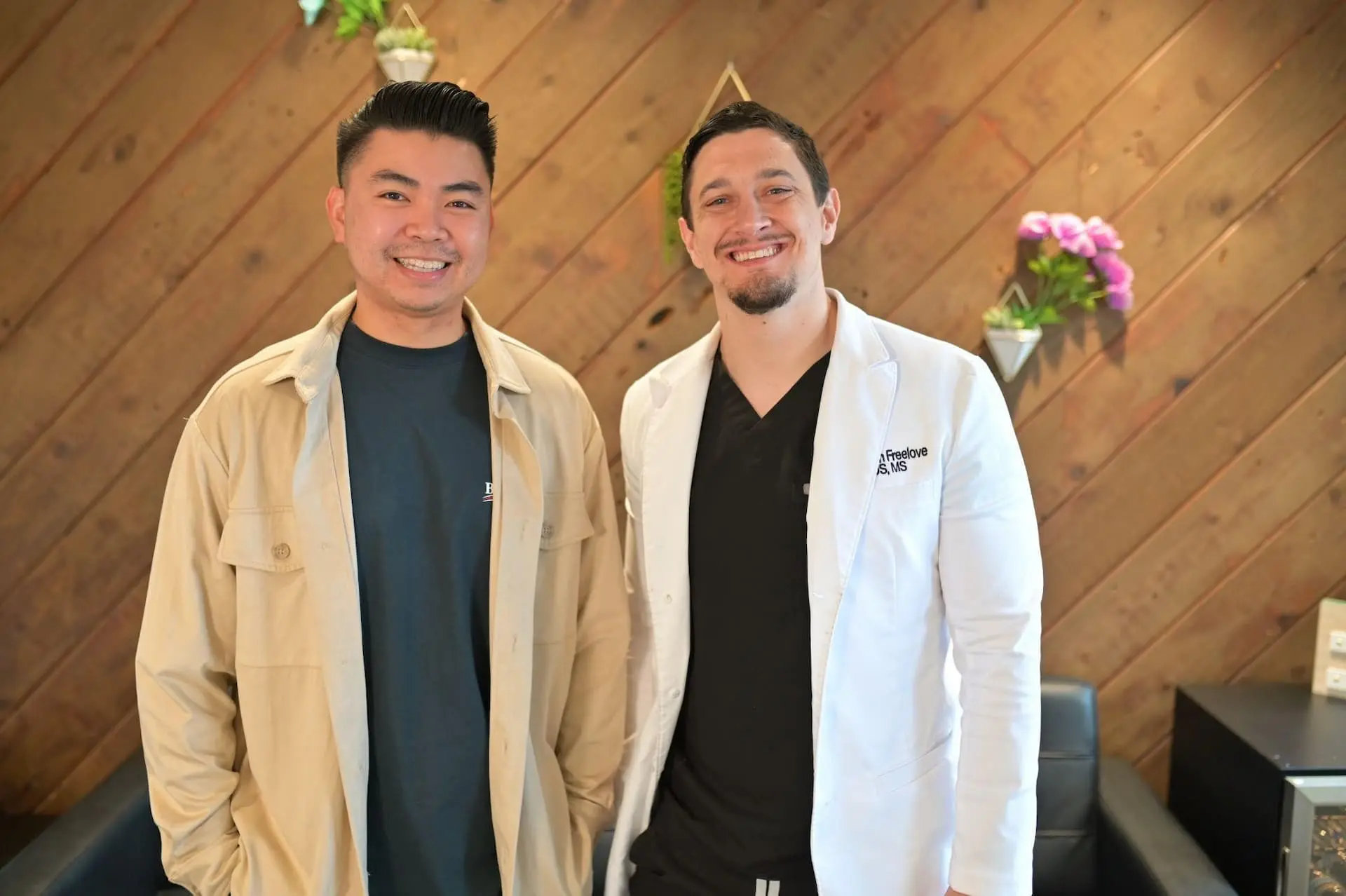 Dr. Cameron Freelove, DDS and his patient smiling in a room with a wooden wall and small shelves with purple flowers in Sammamish WA after her patient's Invisalign clear aligner treatment