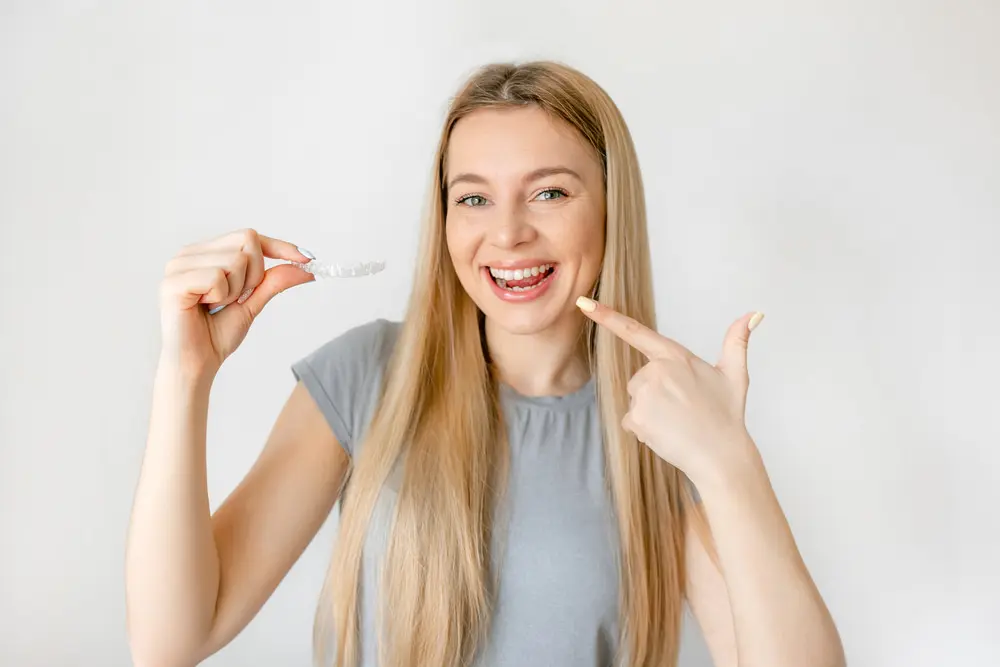 a woman in grey t-shirt pointing at her teeth and holding a Invisalign clear aligner from orthodontist in Sammamish, WA