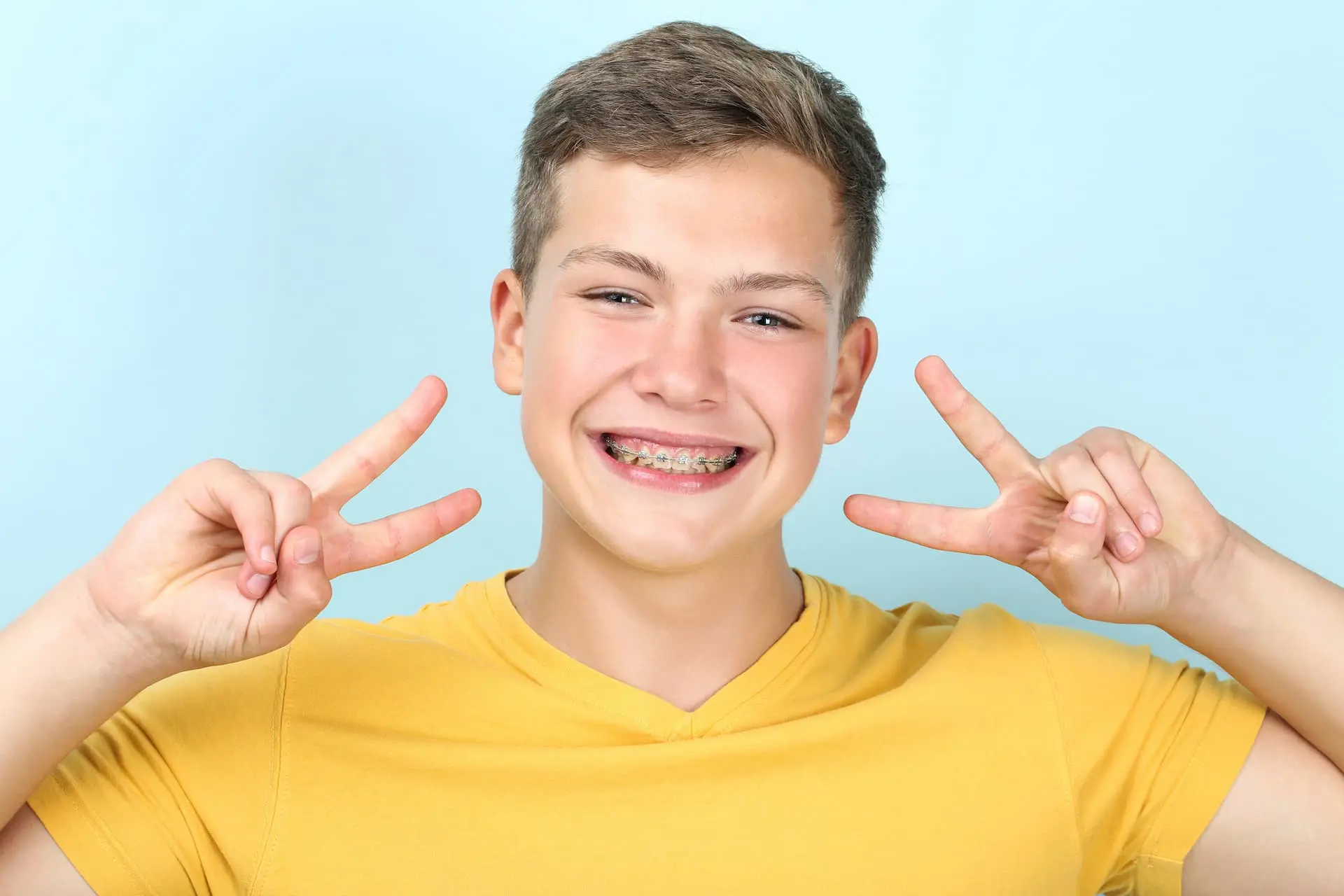 A smiling person wearing a yellow shirt and metal braces, as one of the types of Braces in Sammamish or Kent, WA holds up peace signs with both hands against a blue background.