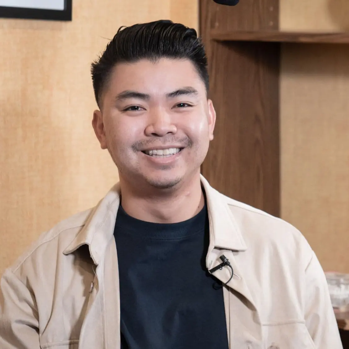 A smiling person with short black hair wears a beige jacket over a black shirt, sitting indoors against a wood-paneled wall at Freelove Orthodontics