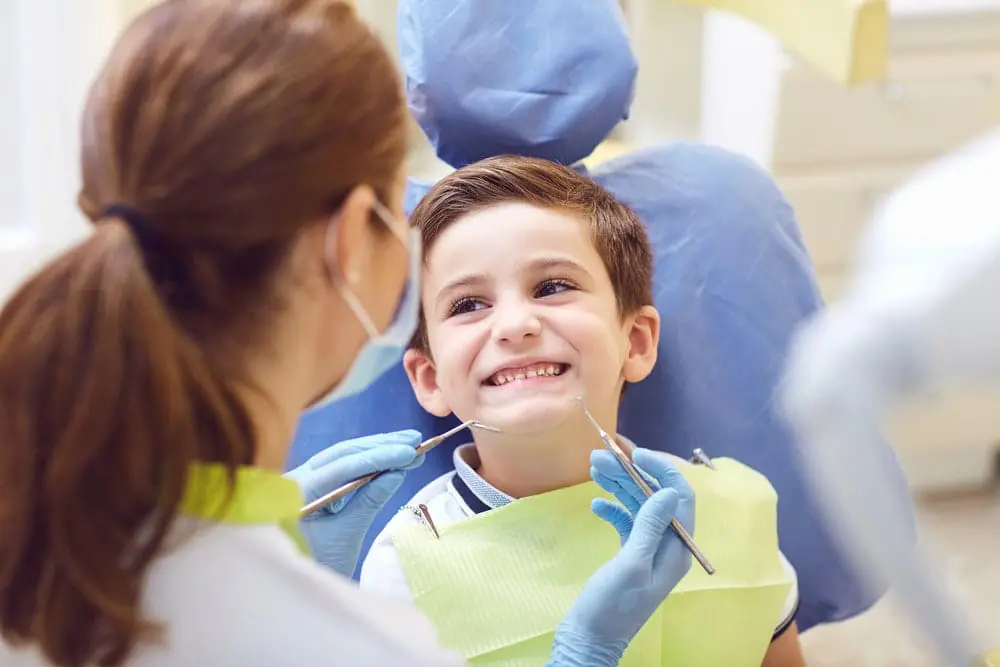 A dentist examines a smiling child in a dental chair. The child wears a green bib, and the dentist holds dental tools. Both are wearing protective masks for his palate expander from orthodontist in Sammamish or Kent, WA.