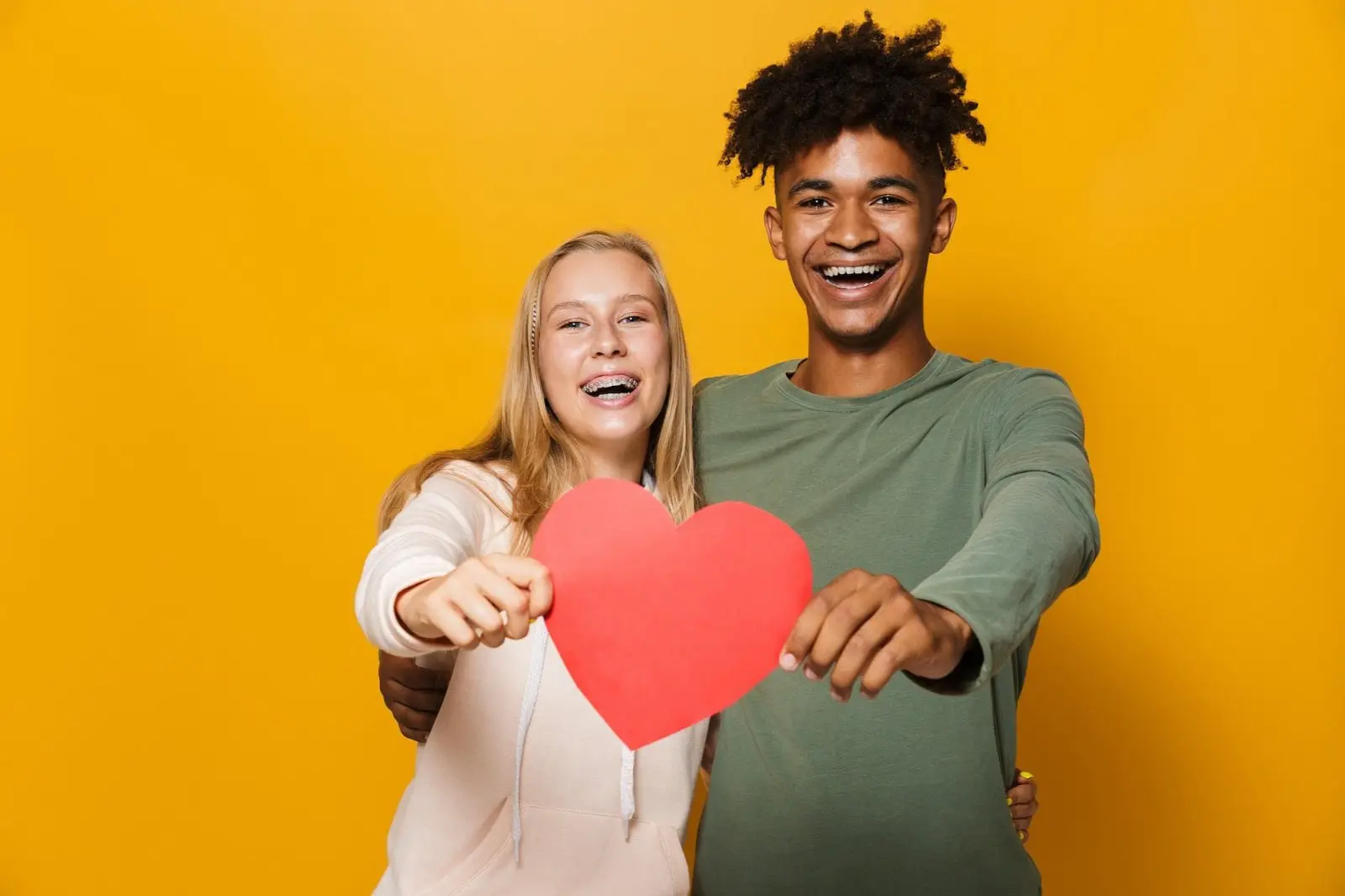 Two people smiling with metal braces from orthodontist in Sammamish or Kent, WA, holding a red heart cutout in front of a vibrant yellow background.