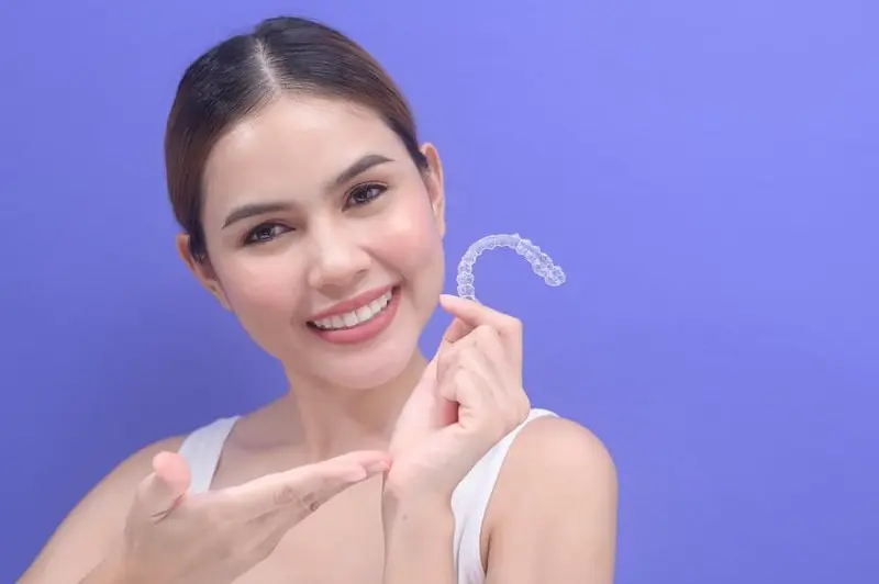 A person smiling and holding a clear dental aligner against a solid purple background, showcasing an alternative to traditional braces covered by orthodontic insurance in Sammamish or Kent, WA.