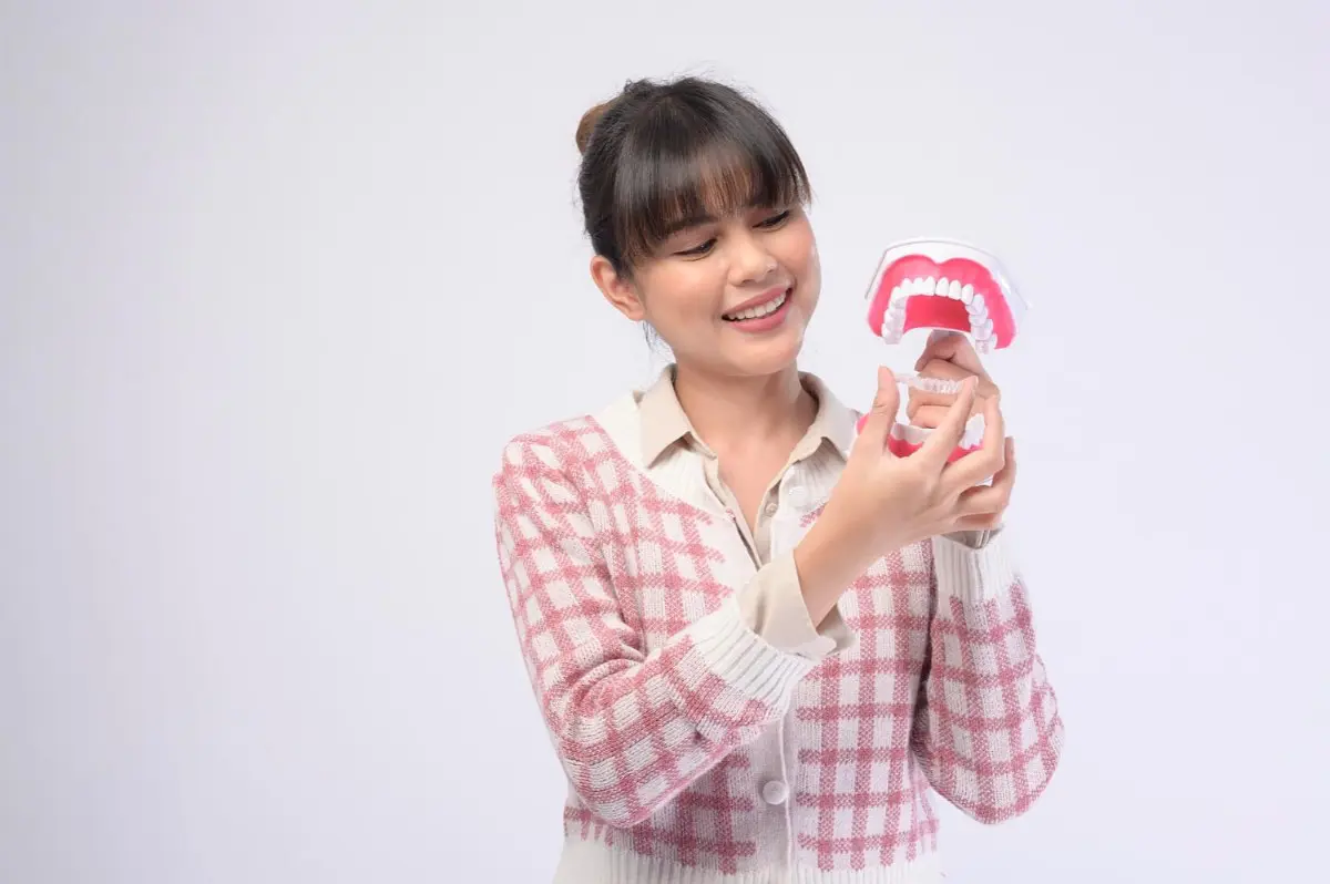 A woman in a pink checkered sweater holds and examines a dental model of teeth, envisioning how an Invisalign clear aligner could transform smiles in Kent, WA, set against a plain background.