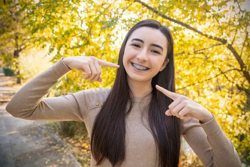 A young woman with long dark hair smiles and points to her Metal Braces in Kent Sammamish, WA braces, standing outdoors in front of trees with autumn leaves.
