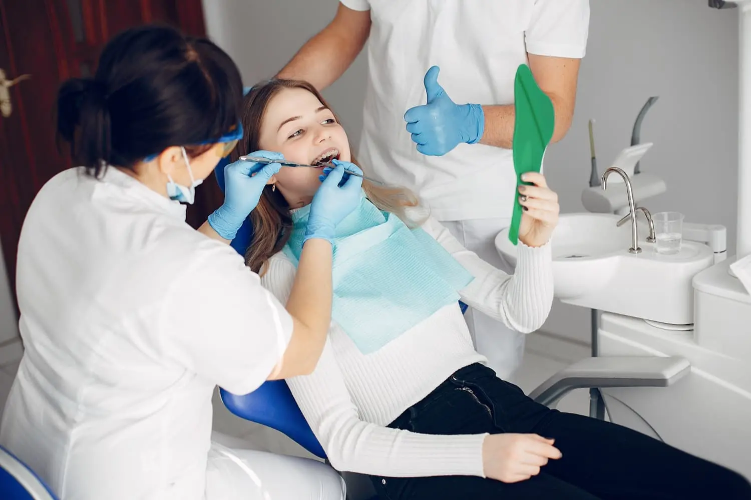 A young woman in a dental chair being fitted with orthodontic appliances at Freelove orthodontics, while holding a green dental mirror. A dental assistant stands nearby.