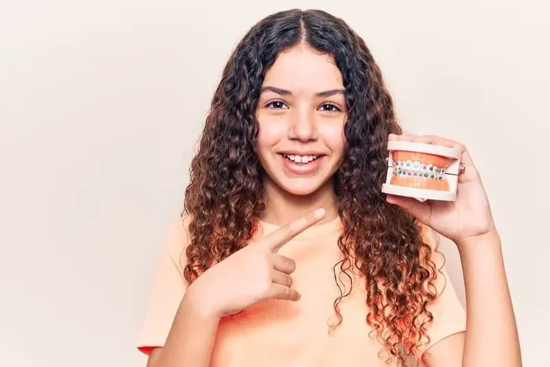 Best Orthodontics Girl with curly hair smiles and points at a dental model with braces, wearing a peach-colored shirt against a plain background. Illustrating Braces Cost Sammamish or Kent, WA