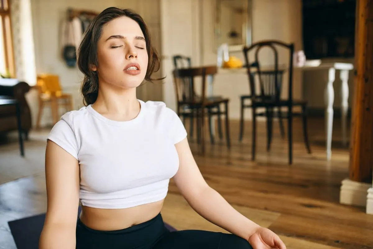 A mouth breather woman in a white shirt sits cross-legged indoors with eyes closed, appearing relaxed. Wooden chairs and a table are visible in the background for orthodontist in Sammamish and Kent, WA.