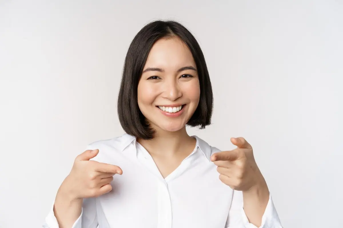 Person with shoulder-length hair, smiling and pointing forward with both hands, showcases a brilliant smile, possibly enhanced by Invisalign clear aligners. Dressed in a white shirt against a white background, this image captures confidence and dental charm often seen around Kent, WA.