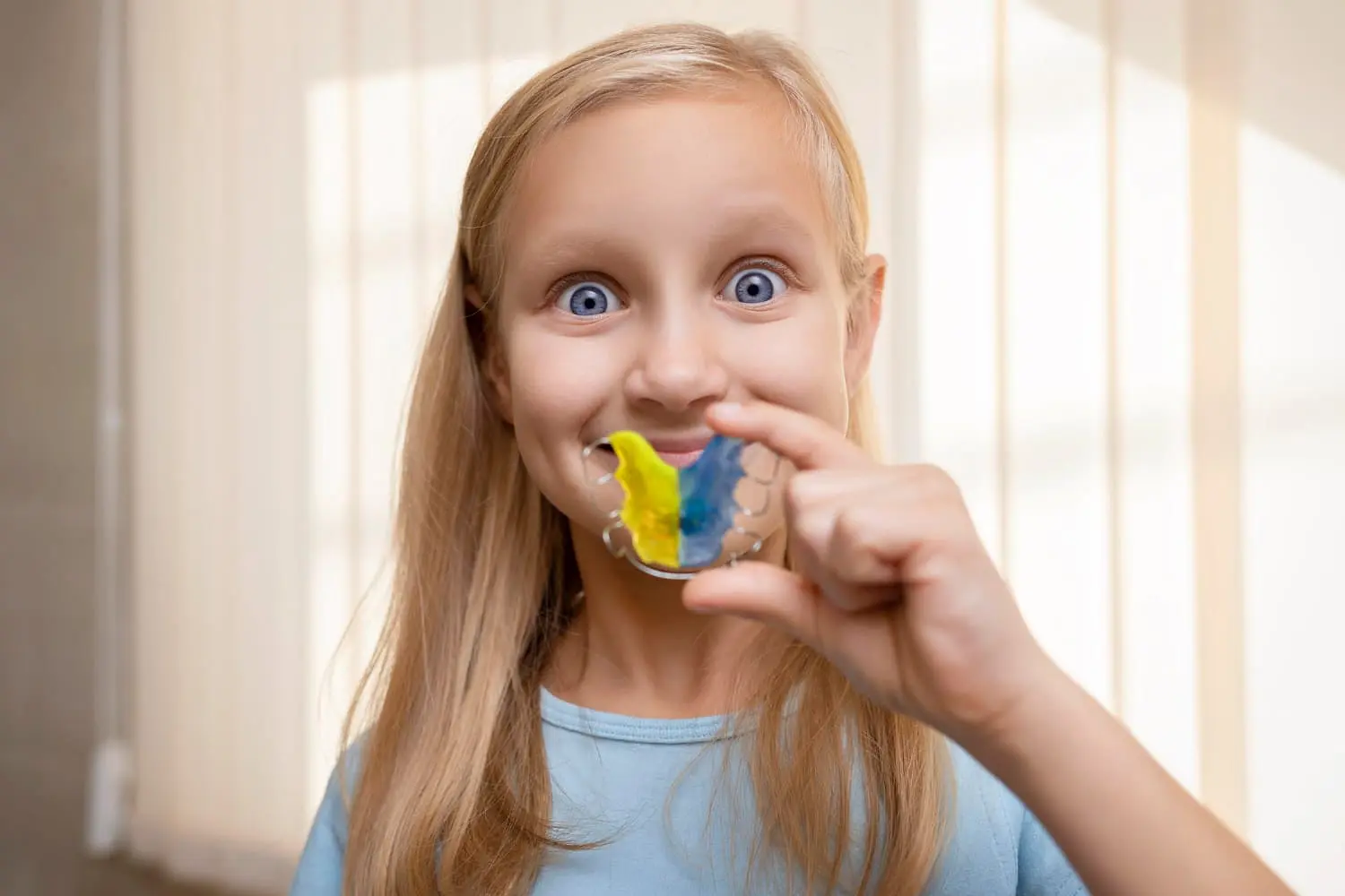 A girl with long blonde hair holds a colorful orthodontic appliances in Kent WA retainer in front of her mouth, smiling widely. She is wearing a light blue shirt in a softly lit room.