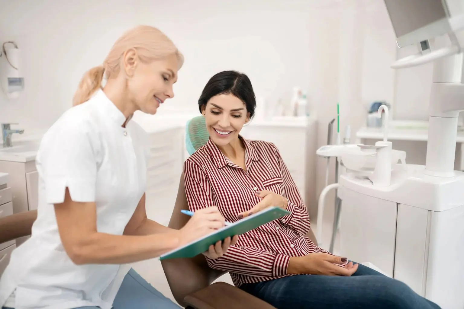 An orthodontist in Sammamish or Kent, WA shows a document on a clipboard to a smiling patient sitting for an affordable cheap Invisalign in a dental chair in a modern clinic.