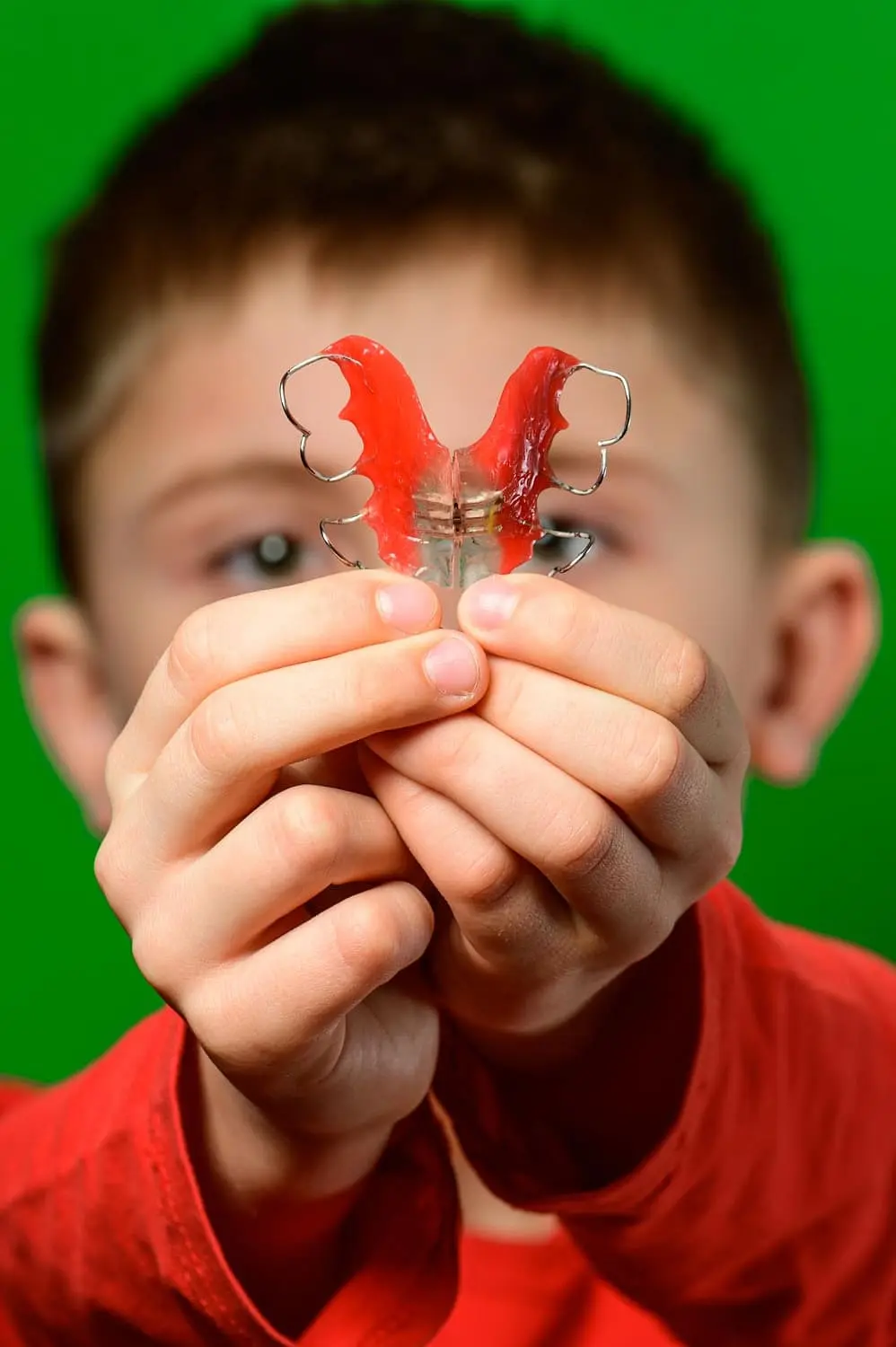 Child holding a red orthodontic appliances sammamish wa retainer with both hands against a green background.