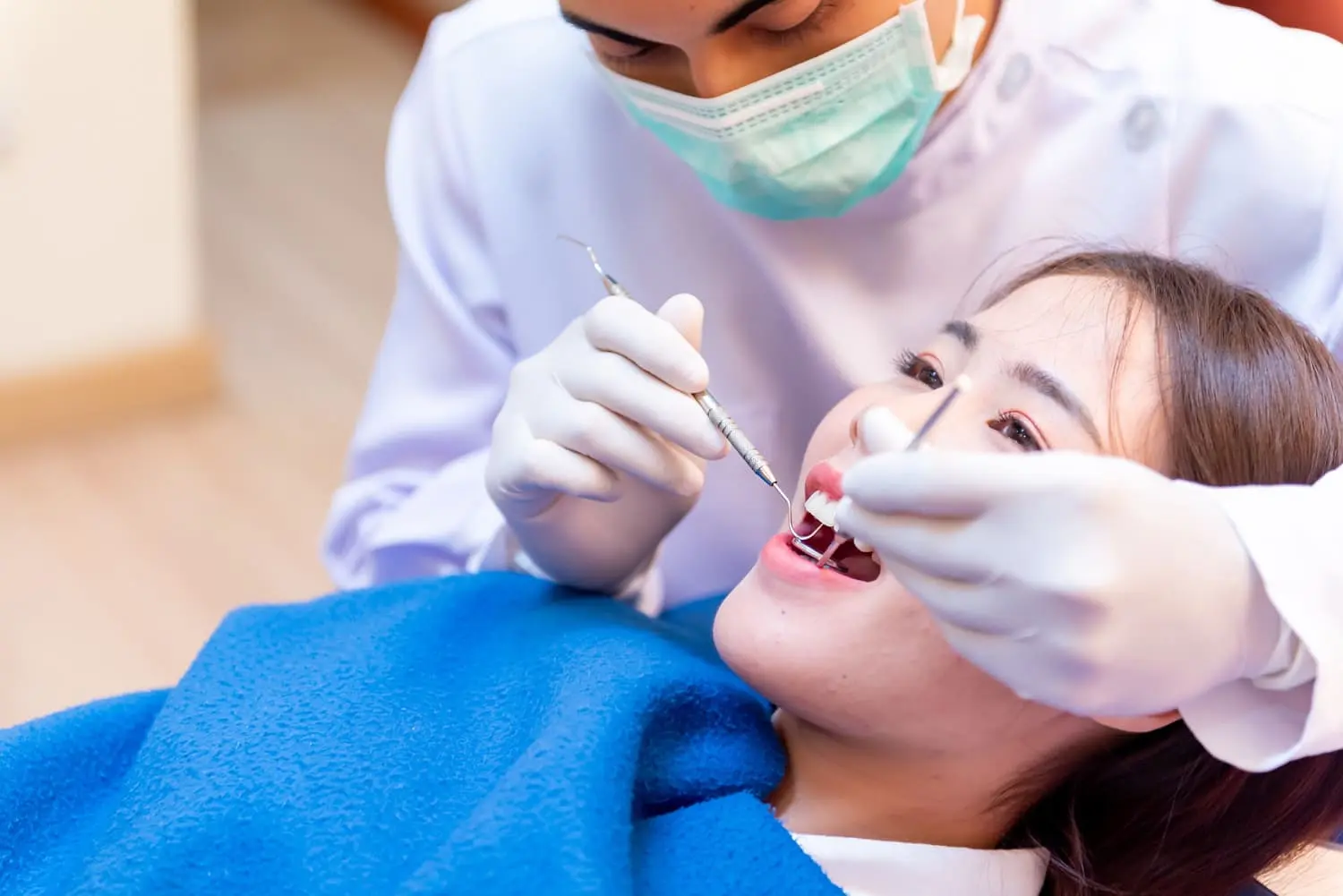 Orthodontist wearing a mask and gloves examines a patient's teeth with orthodontic appliances in Sammamish, WA while the patient lies in a chair, covered with a blue blanket.