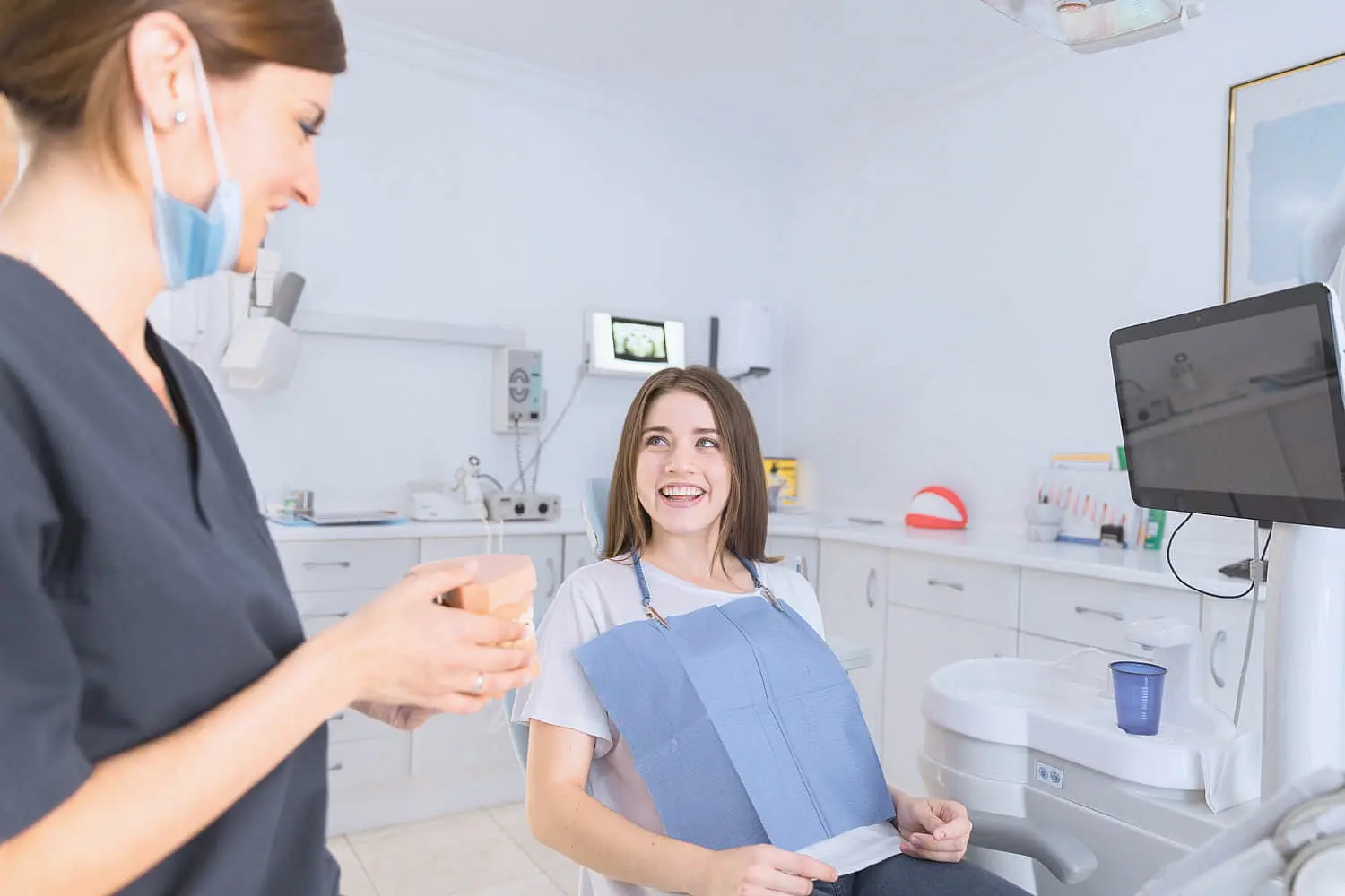 Dentist and patient smiling at each other in a dental clinic. The patient sits in a dental chair with a bib, while the dentist holds a small cup in Sammamish or Kent, WA.