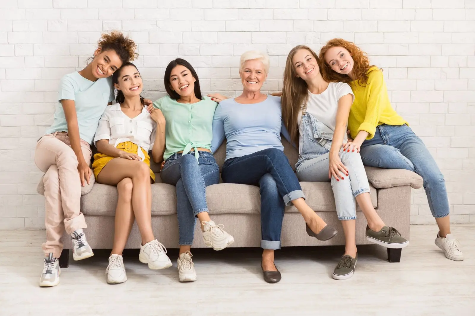 Six women of various ages and ethnic backgrounds sit on a couch, smiling at the camera against a white brick wall, perhaps discussing the benefits of an Invisalign payment plan.