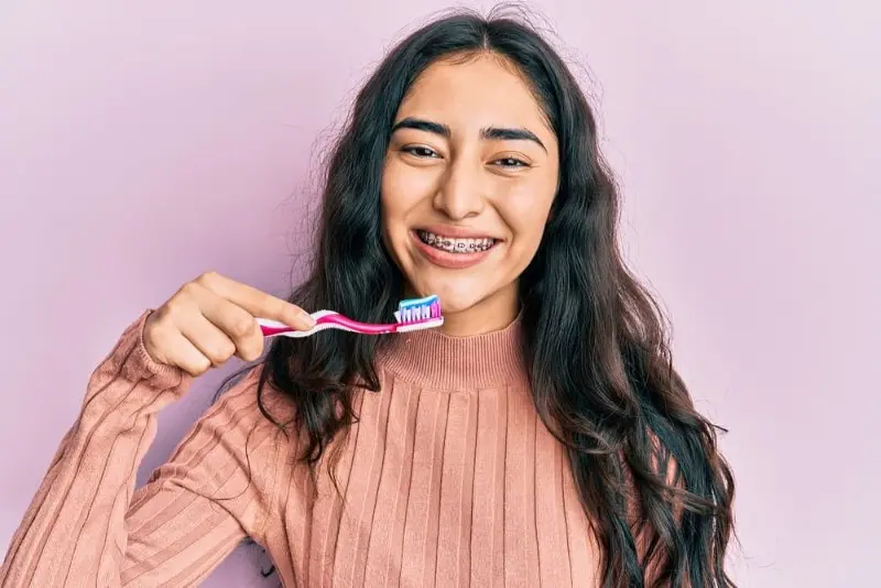 A person with long hair and metal braces stands against a pink background in Kent, WA, holding a toothbrush and smiling widely.