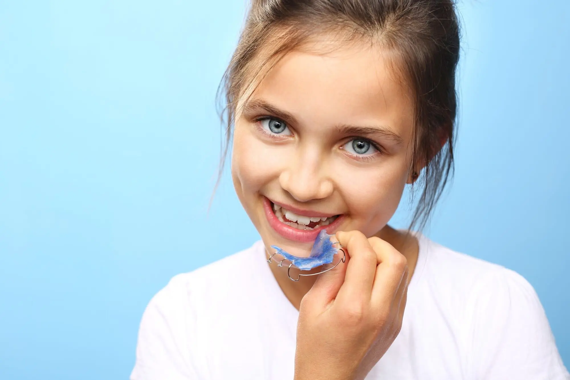 A girl with blue eyes and brown hair smiles while holding a blue dental retainer near her mouth, against a light blue background.