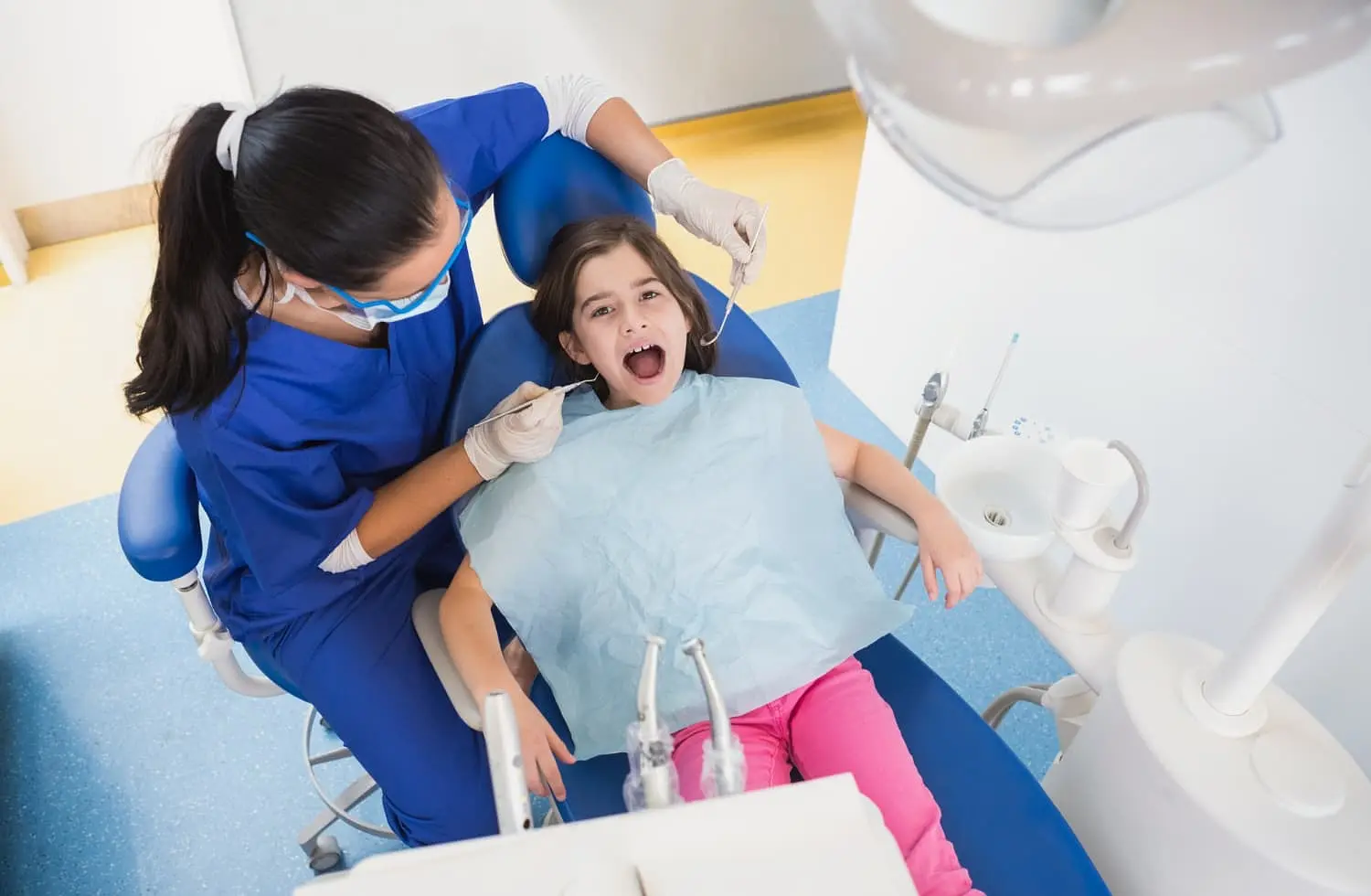 A dentist examines a young girl with orthodontic appliances in kent wa sitting in a dental chair with her mouth open. The dentist wears blue scrubs, gloves, and a mask. The girl wears a bib and pink pants. Dental tools are visible.