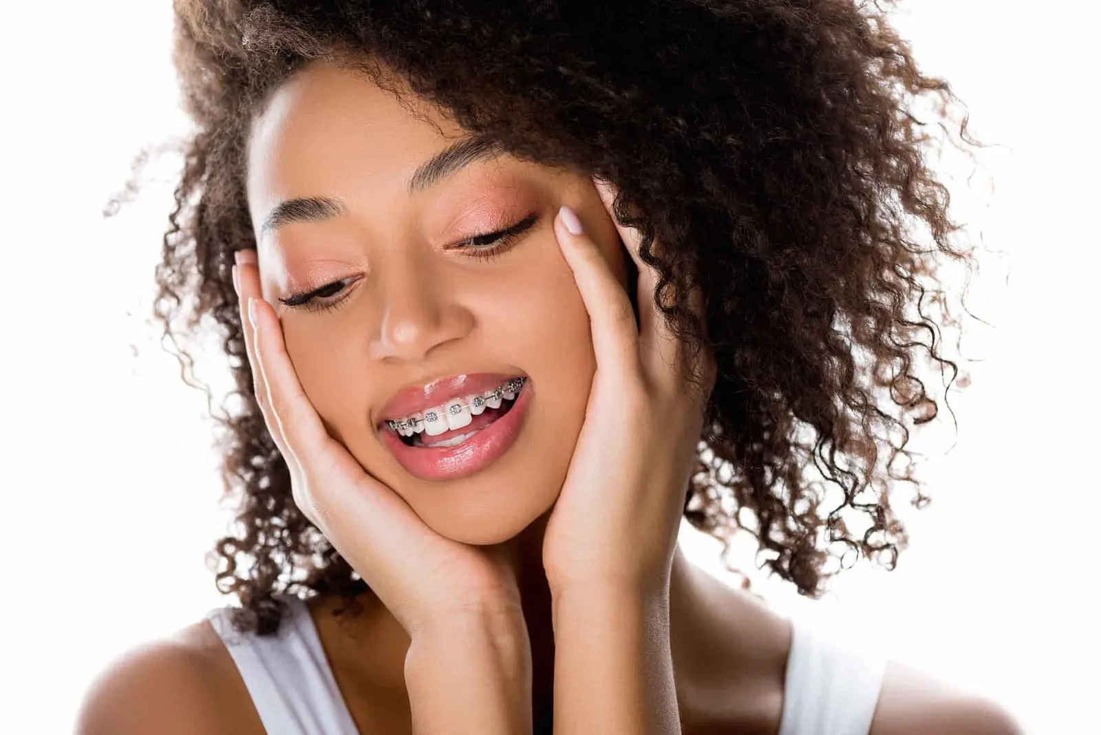 Person with curly hair and braces smiling, hands touching cheeks, eyes closed, against a white background. Illustrating Types of Braces Sammamish or Kent, WA