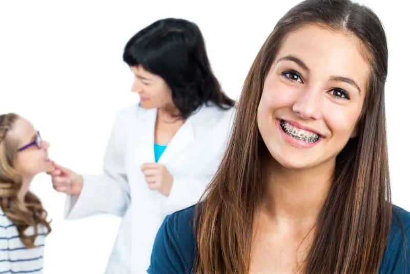 Best Orthodontics Teen girl with braces smiles at the camera; in the background, a woman in a white coat examines a young girl wearing glasses. Illustrating Braces Cost Sammamish or Kent, WA