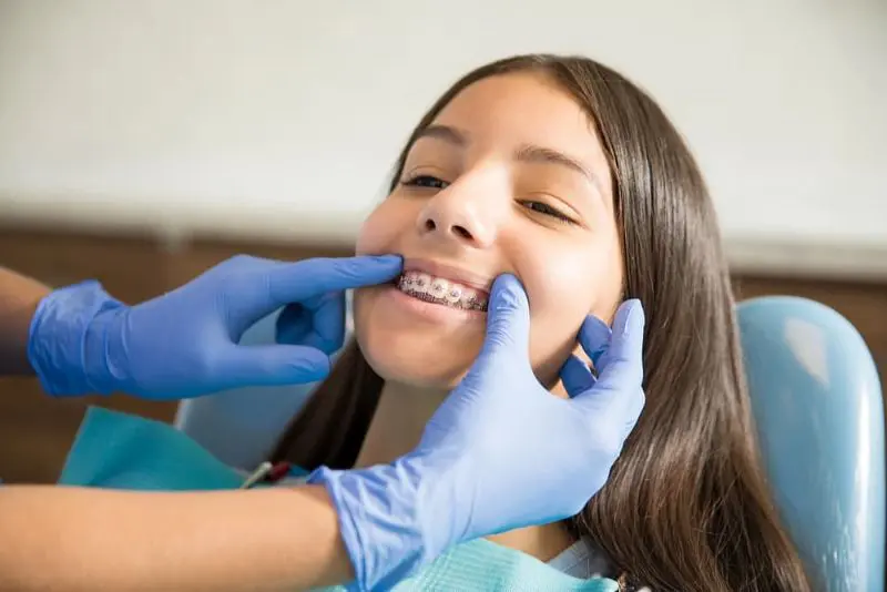 Best Orthodontics A dentist wearing blue gloves adjusts a girl's braces as she sits in a dental chair, smiling slightly. Illustrating Braces Cost Sammamish or Kent, WA