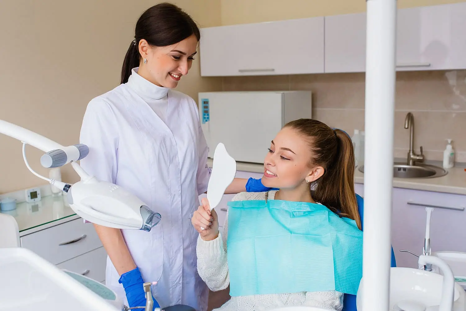 Dentist in a white coat and gloves stands beside a seated patient holding a dental mirror. The patient wears a blue bib and smiles, holding the mirror to view her teeth in Sammamish, WA.