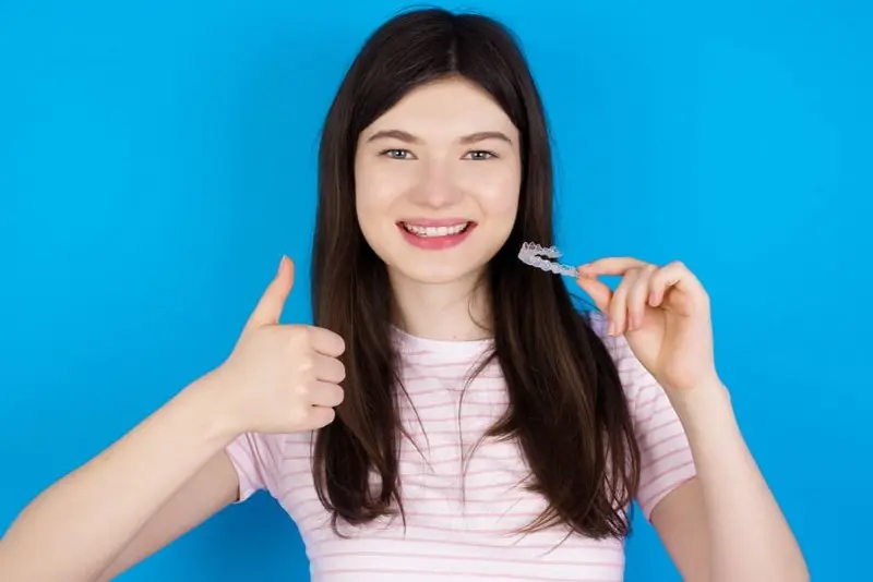 Young woman smiling, holding a clear dental aligner in one hand, giving a thumbs-up with the other, standing against a blue background. Her expression suggests she's thrilled with her choice in the Invisalign vs braces debate in Sammamish and Kent, WA.
