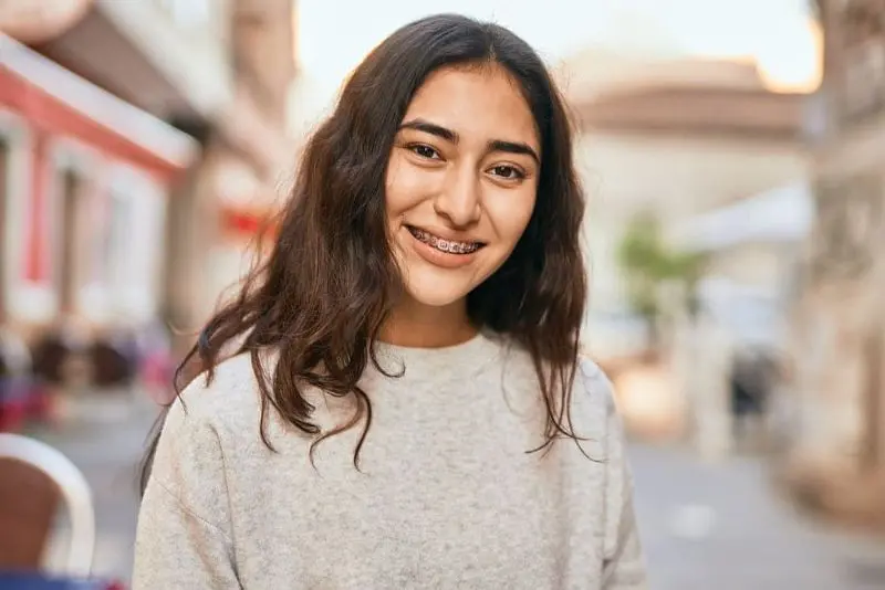 A woman with long dark hair and braces smiles while standing outdoors on a city street. She is wearing a light gray sweater. Illustrating Metal Braces in Kent Sammamish, WA