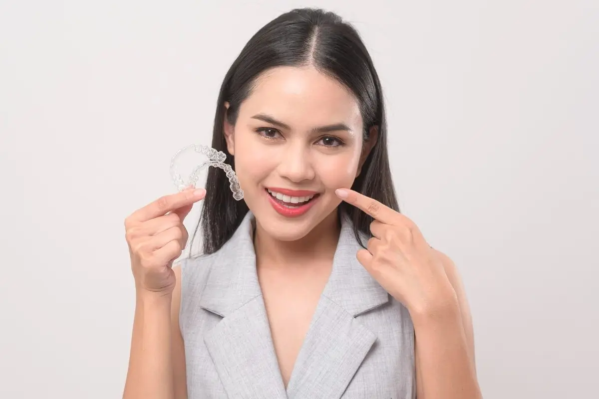 A woman smiles while holding a clear dental aligner in one hand and pointing to her teeth with the other, proudly showcasing her journey with adult braces from orthodontist in Sammamish or Kent, WA. She is wearing a light gray sleeveless top.