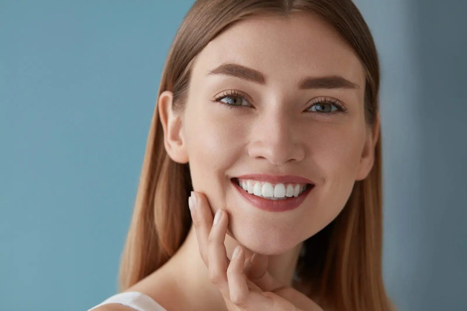 A woman with long hair smiles, touching her face against a blue background, reminiscent of the serene skies near orthodontist in Sammamish or Kent, WA.