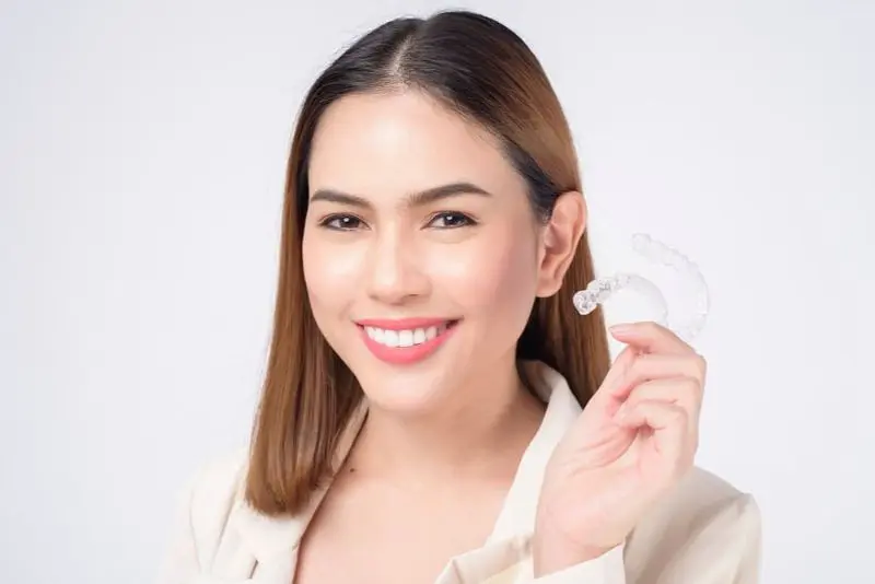A woman smiles brightly, holding a clear dental aligner in her right hand against a plain white background, showcasing her confidence with Invisalign and the flexibility of their payment plan in Sammamish or Kent, WA.