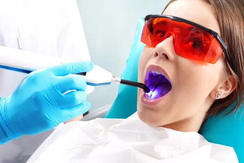 A dental professional uses a blue curing light on a patient's teeth. The patient wears red protective glasses. Illustrating Invisalign vs braces in Sammamish or Kent, WA.