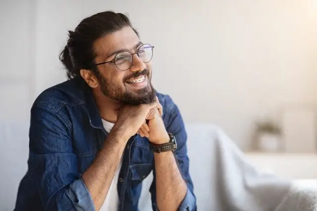 A man with glasses and a beard smiles while resting his chin on his hands, exuding the friendly vibe near orthodontist in Sammamish or Kent, WA. He is wearing a denim shirt and sitting in a bright room.