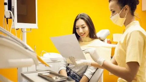 A dental professional in a mask shows a document to a seated woman in a dental office with bright yellow walls. Illustrating braces orthodontic insurance Sammamish or Kent, WA