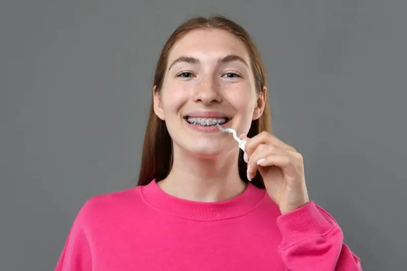 A person in a pink sweater, smiling and holding a dental flosser near their braces, against a gray background. Illustrating Invisalign vs braces in Sammamish or Kent, WA