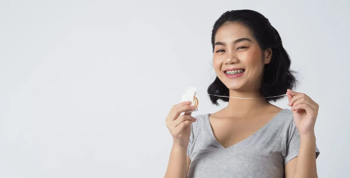 A woman with braces smiles while holding dental floss, standing against a plain background. Illustrating Metal Braces in Kent Sammamish, WA