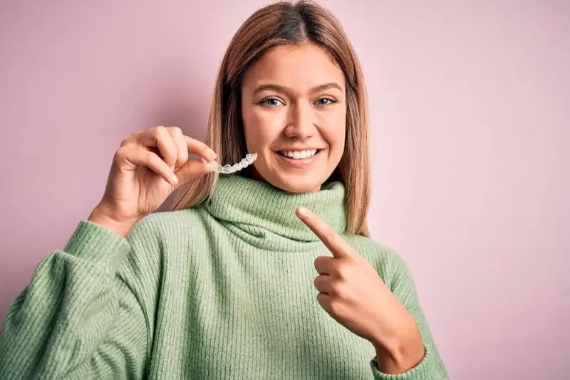 Person in a green sweater holds and points to a Invisalign dental aligner showing Invisalign Payment Plan, standing against a pink background.
