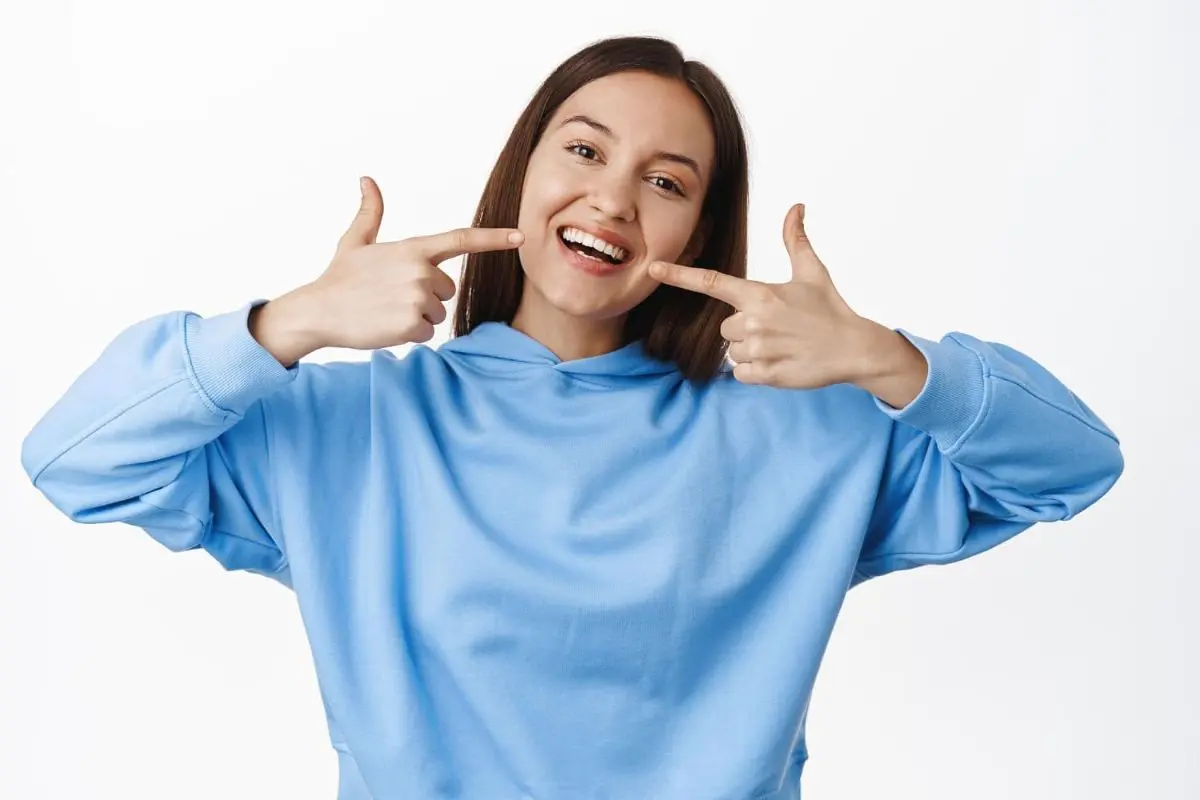 Person in a blue hoodie pointing at their smile with Metal Braces in Kent Sammamish, WA with both hands against a plain background.