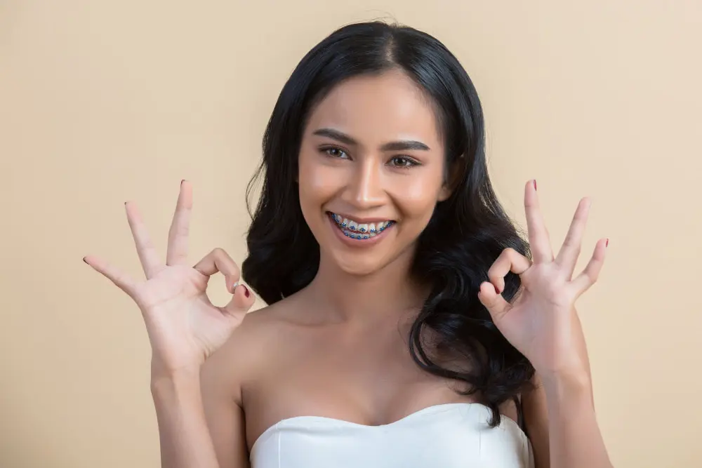 A woman with long dark hair and braces is smiling, wearing a strapless white top, and making an "OK" gesture with both hands against a beige background. Illustrating adult braces in Sammamish and Kent, WA