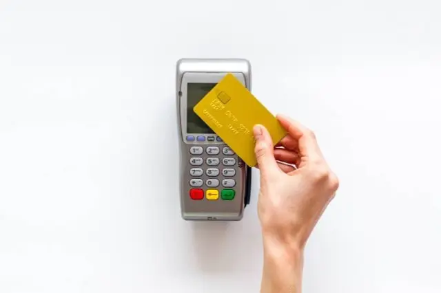 A hand holds a yellow credit card over a payment terminal with buttons and a display screen, ready to settle an braces and orthodontic insurance charge in Sammamish or Kent, WA, all set against a white background.
